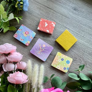 May include: An overhead shot displays five small, square, patterned paper notebooks. The notebooks feature floral, geometric, and nature-inspired designs in various colors, including pink, blue, yellow, and purple. The notebooks are arranged on a wooden surface.
