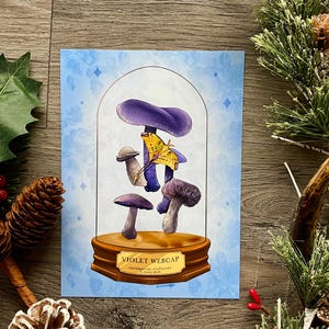 May include: A print featuring a stylized illustration of violet webcap mushrooms under a glass dome. The artwork includes a yellow butterfly and the text "VIOLET WEBCAP" on a wooden base, set against a light blue background.
