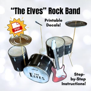 Puede incluir: Un set de batería de banda de rock DIY con tambores, una guitarra y un micrófono. Los tambores están hechos de recipientes negros y plateados, y la guitarra es roja. El set incluye el texto "The Elves" y "Printable Decals!"