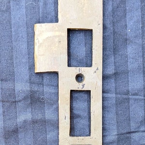 May include: A rectangular brass door strike plate with three rectangular cutouts and two screws. The plate has a slightly tarnished appearance and is lying on a blue striped fabric.