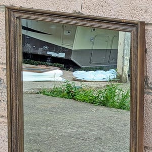 May include: A tall, rectangular mirror with a brown wooden frame. The frame has a decorative rope-like texture around the inner edge. The mirror reflects the surrounding environment, including a vehicle and greenery.