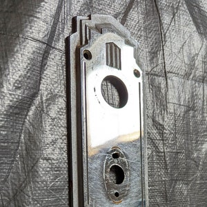May include: A silver door plate with an Art Deco design. The plate has a keyhole and several decorative lines.
