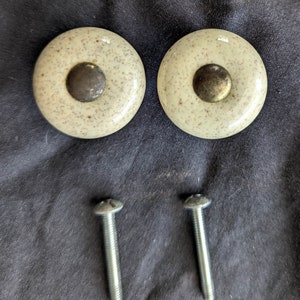 May include: Two beige ceramic knobs with a speckled finish and a gold metal center. Two silver screws are included.