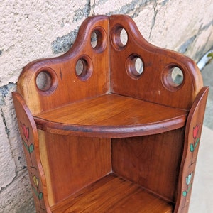May include: A wooden corner shelf with three tiers. The shelf is painted with a floral design featuring tulips in red, yellow, and green. The shelf has a rounded top and a decorative design with holes.
