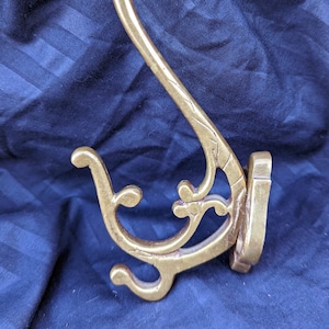 May include: A gold-toned metal hook with a decorative, ornate design. The hook is curved and has a flat base.