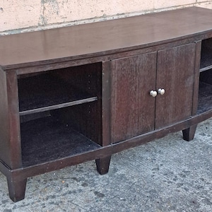 May include: A dark brown wooden media console with two doors and open shelving. The cabinet has a curved front and sits on short, square legs. The doors have silver knobs.