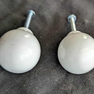 Może przedstawiać: Dwa białe ceramiczne gałki do szafek ze srebrnymi metalowymi śrubami. Gałki są okrągłe i mają gładkie, błyszczące wykończenie.