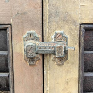 May include: Close-up of a vintage metal door latch with an aged, weathered appearance. The latch is mounted on two wooden door panels with visible grain and a distressed finish. The latch has decorative detailing.