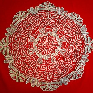 May include: A white crocheted lace doily with a circular design featuring intricate floral patterns and a central motif. The doily is displayed on a red background.
