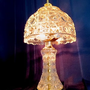 May include: A table lamp featuring a clear, cut-glass dome shade and a matching cut-glass base. The lamp has a polished gold-tone metal finial at the top, a gold-tone metal neck, and a circular gold-tone metal base with an attached power cord.