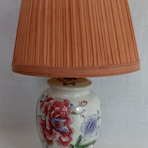 May include: A vintage table lamp with a peach-colored pleated lampshade. The white ceramic base features hand-painted floral designs in red, pink, and blue. The lamp has a dark red base and a two-prong plug.