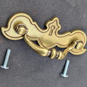 Könnte beinhalten: Ein goldfarbener Metall-Schrankgriff mit einem dekorativen, verzierten Design. Der Griff hat ein Schlüssellochdetail und zwei Schrauben zur Montage.