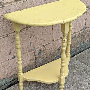 May include: A small, half-moon shaped, yellow side table with a lower shelf. The table has turned legs and a distressed finish, showing signs of wear. The table is approximately 30 inches long.