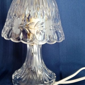May include: A clear glass table lamp featuring a bell-shaped shade with a scalloped edge and etched floral patterns. The base is fluted glass with a brass-colored finial at the top. A white electrical cord with a thumb switch is attached.