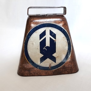 May include: A vintage copper cowbell with a blue and white logo featuring a stylized eagle design. The bell has a worn, rustic look.