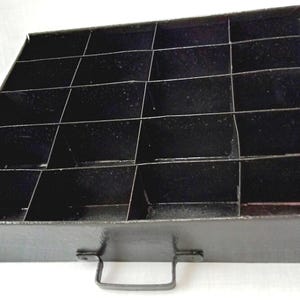 May include: Black metal tray with multiple compartments, ideal for organizing small items. Features a handle for easy carrying. The tray is approximately 12 inches wide and 12 inches long.