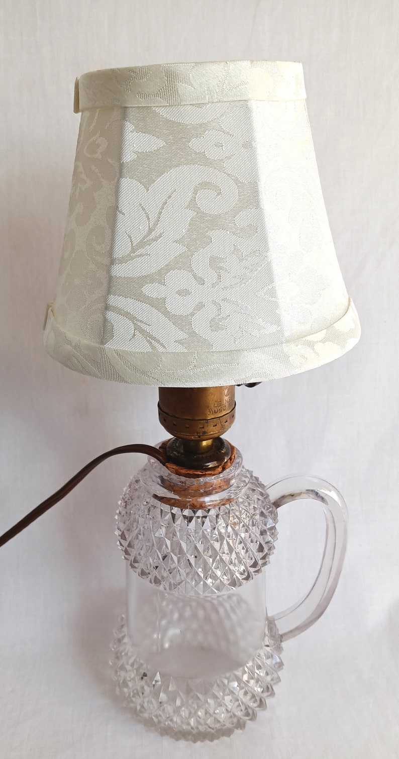 May include: A white lampshade with a floral pattern sits atop a clear glass lamp base with a diamond-shaped pattern. The lamp base has a handle and a gold-coloured metal socket.