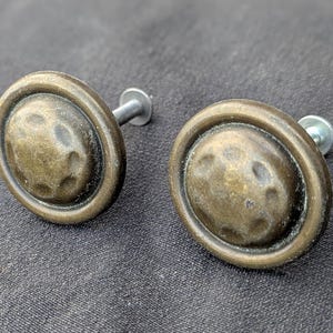Puede incluir: Dos tiradores de armario de latón antiguo con textura martillada sobre fondo oscuro. Cada tirador tiene un diseño circular con centro en relieve y tornillo de fijación. Ideales para proyectos de restauración de muebles.