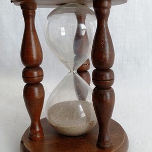 May include: An hourglass with a wooden frame and base. The clear glass bulb contains sand, and the wooden frame is a dark brown color. The hourglass is a decorative item, and the sand is flowing from the top bulb to the bottom.