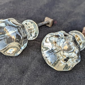 Puede incluir: Dos pomos de puerta de vidrio transparente con superficies facetadas y tallos metálicos. Los pomos están sobre un fondo gris oscuro.