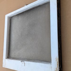 May include: An antique window frame with a textured glass pane. The frame is painted white with visible wear and tear, and the edges are dark brown. A small latch is visible on the bottom of the frame.