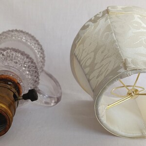 May include: A clear glass lamp base with a textured surface and a brass light socket. A white fabric lampshade with a floral pattern.