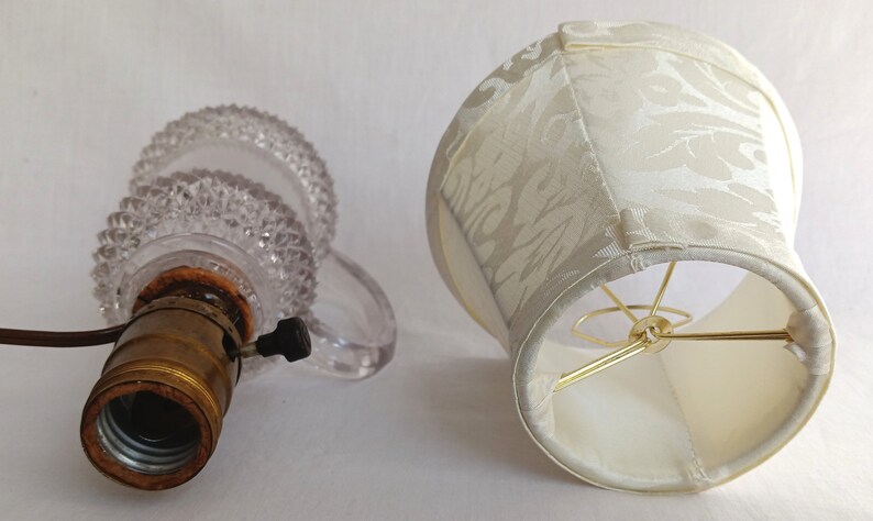 May include: A clear glass lamp base with a textured surface and a brass light socket. A white fabric lampshade with a floral pattern.