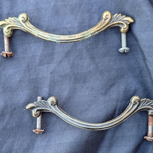 May include: Two antique brass drawer pulls with ornate, curved designs. Each pull has a decorative flourish at the ends and includes screws for mounting. The pulls have a vintage, aged appearance.