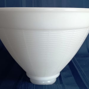May include: A white, cone-shaped glass lampshade with a ribbed texture. The lampshade has a wide top and a smaller, rounded base. The overall shape is reminiscent of a vintage design.