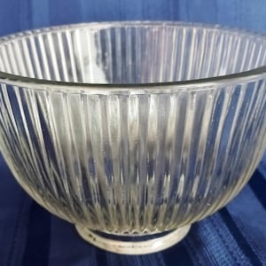 May include: Clear glass bowl with vertical ribbed design. The bowl has a rounded shape with a wide opening and a small, circular base. The glass is transparent, allowing light to pass through, and the ribbed texture adds visual interest.