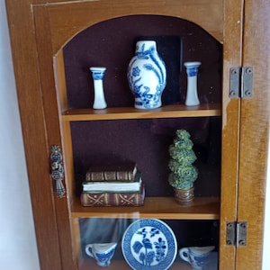 May include: A miniature wooden display cabinet with glass doors. Inside are shelves holding small decorative items: a blue and white vase, two small candlesticks, a stack of books, a topiary, a plate, and two teacups. The cabinet is made of wood.