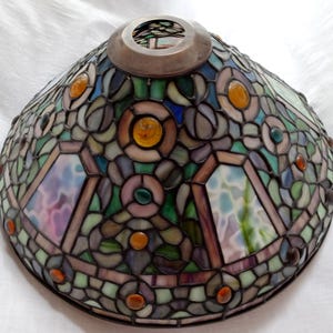 May include: A stained glass lampshade with a cone shape. The shade features a mosaic of colorful glass pieces in shades of green, blue, purple, and orange. The design includes floral and geometric patterns, with a silver metal ring at the top.