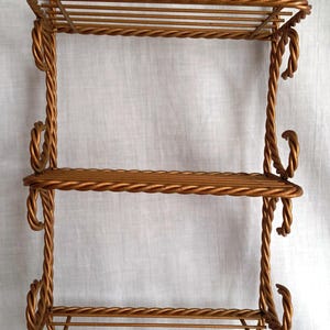 May include: A decorative, three-tiered, gold-colored metal wall shelf. The shelf has a twisted rope design on the frame and shelves. The top has a circular loop for hanging. The bottom has two decorative tassels.
