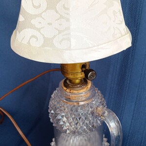 May include: A vintage table lamp with a clear glass base featuring a diamond pattern and a white fabric lampshade with a floral design. The lamp has a brown cord and a black switch.