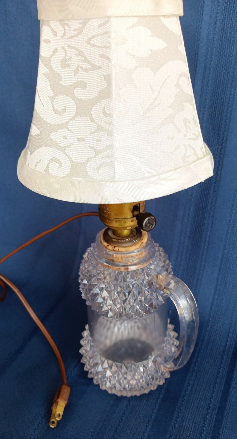 May include: A vintage table lamp with a clear glass base featuring a diamond pattern and a white fabric lampshade with a floral design. The lamp has a brown cord and a black switch.