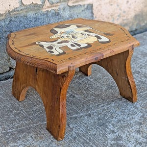 May include: A wooden step stool with a painted design of two cows wearing hats. The stool is brown and has a rounded top.