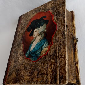 May include: A vintage wooden box with a hinged lid. The front features a framed portrait of a woman in a blue dress and a black hat. The box has a distressed, wood-grain finish, and a red border surrounds the portrait.