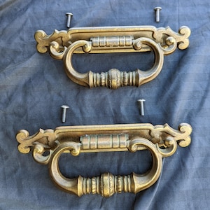 May include: Two antique brass drawer pulls with ornate scrollwork and a curved handle design. Each pull includes two screws for mounting. The pulls have a warm, aged patina, suggesting vintage craftsmanship. Ideal for furniture restoration or adding a classic touch.