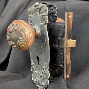 Könnte beinhalten: Antikes Türbeschlagsset mit einem runden, kupferfarbenen Türknauf mit einem verzierten Design. Das Set enthält eine schwarze Metallplatte mit dekorativen Verzierungen und einen messingfarbenen Riegel. Für eine Tür.