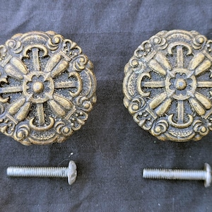 May include: Two antique brass-colored cabinet knobs with ornate floral designs. Each knob has a central flower shape surrounded by detailed scrollwork and a matching screw for installation. The knobs are approximately 2 inches in diameter.