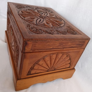 May include: A wooden box with intricate carvings. The box is a warm brown color and features detailed floral and geometric patterns on the lid and sides. The box sits on a small, raised base.