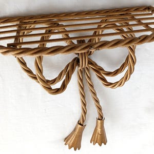 May include: A decorative wall shelf made of twisted rope-like material. The shelf has a rectangular shape with a bow design and two tassel-like ends. The color is a warm, light brown.