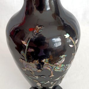 May include: Black decorative vase with a glossy finish and a gold rim. The vase features iridescent floral and leaf designs inlaid on the surface. The vase has a wide body and a narrow neck, and sits on a round base.