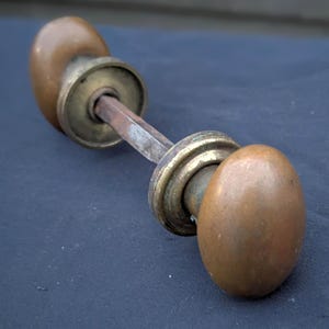 May include: Antique door handle with two oval-shaped bronze knobs and a connecting rod. The handle has a weathered appearance, with signs of age and use. The metal has a warm, brown patina.
