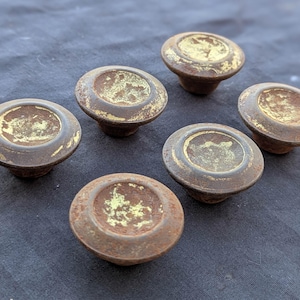 Könnte beinhalten: Sechs antike Schrankknöpfe mit einem abgenutzten, gealterten Aussehen. Die Knöpfe sind hauptsächlich braun mit einer goldfarbenen Mitte und weisen Gebrauchsspuren und Rost auf. Diese Vintage-Knöpfe eignen sich für Möbelrestaurierungsprojekte.