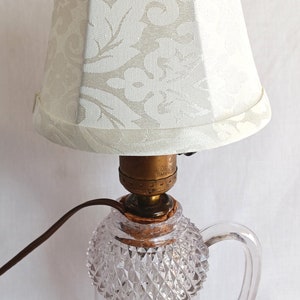 May include: A white lampshade with a floral pattern sits atop a clear glass lamp base with a diamond-shaped pattern. The lamp base has a handle and a gold-coloured metal socket.