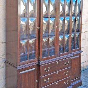 May include: A dark wood display cabinet with glass-paned doors and three drawers. The cabinet has a rectangular shape with a decorative top and brass hardware. The glass reflects the surrounding environment, adding depth to the design.