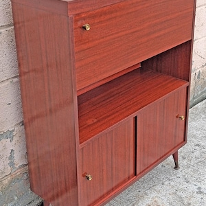 May include: A tall, rectangular wooden cabinet with a rich reddish-brown wood grain finish. It features a top drop-down door, an open middle shelf, and two lower sliding doors. Each door has a small, circular brass knob. The unit stands on four tapered legs with brass-toned metal feet.