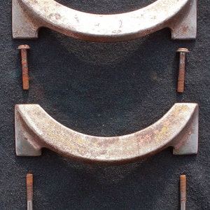 Op de afbeelding: Twee roestige metalen handvatten met afgeronde randen en schroefgaten. Elk handvat is bevestigd aan een roestige schroef.