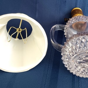 May include: A white lampshade with a gold metal harp and a clear glass lamp base with a diamond pattern.